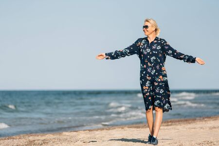 End of quarantine. Young blonde woman in sunglasses and long dress walking on beachの写真素材