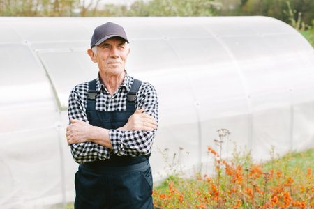 Smiling cheerful senior gardener standing in front of a greenhouse. Old man in cap and overall in...
