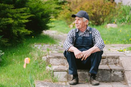 Senior gardener sitting on the steps in garden. Aged worker in overall and baseball cap resting after work. Portrait of old farmerの写真素材