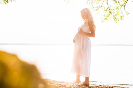 Pregnant blonde woman in long white dress on the beach. Future mother with bellyの写真素材