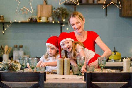 Mother with Two Happy cheerful Little girls in santa hat sitting at the festive table. Christmas...