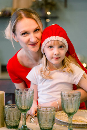 Mother with Happy cheerful Little girl in santa hat sitting at the festive table. Christmas time. Woman having fun with daughterの写真素材