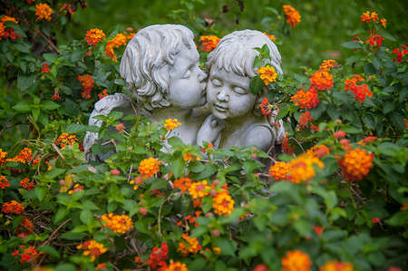 ANGELS IN LOVE. Sculptures of boy and girl angels surrounded with flowers hugging and cuddling. The boy angel is kissing her by the cheek.の写真素材