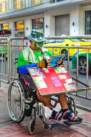 China Town, Bangkok, Thailand - Nov. 14, 2020: Everyone wears protective facemask. A lottery seller on wheelchair wearing fancy protective facemask gazing on his tablet while waiting for customers.のeditorial素材