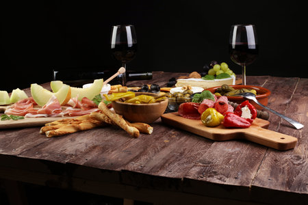 Italian antipasti wine snacks set. Cheese variety, Mediterranean olives, pickles, Prosciutto di Parma with melon, salami and wine in glasses over black grunge backgroundの写真素材