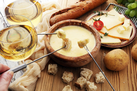 Gourmet Swiss fondue dinner on a winter evening with assorted cheeses on a board alongside a heated pot of cheese fondue with two forks dipping bread and white wine behind in a tavernの写真素材