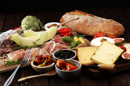 Italian antipasti wine snacks set. Cheese variety, Mediterranean olives, pickles, Prosciutto di Parma with melon, salami and wine in glasses over black grunge backgroundの写真素材