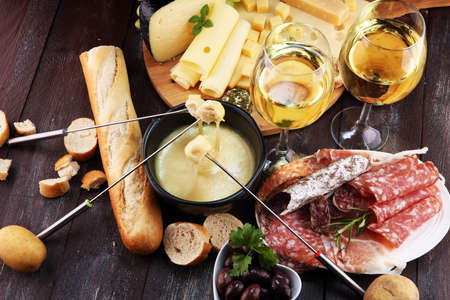 Gourmet Swiss fondue dinner on a winter evening with assorted cheeses on a board alongside a heated pot of cheese fondue with two forks dipping bread and white wine behind in a tavern or restaurantの写真素材