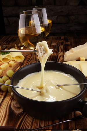 Gourmet Swiss fondue dinner on a winter evening with assorted cheeses on a board alongside a heated pot of cheese fondue with two forks dipping bread and white wine behind in a tavern or restaurantの写真素材
