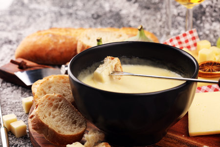 Gourmet Swiss fondue dinner on a winter evening with assorted cheeses on a board alongside a heated pot of cheese fondue with two forks dipping bread and white wine behind in a tavern or restaurantの写真素材