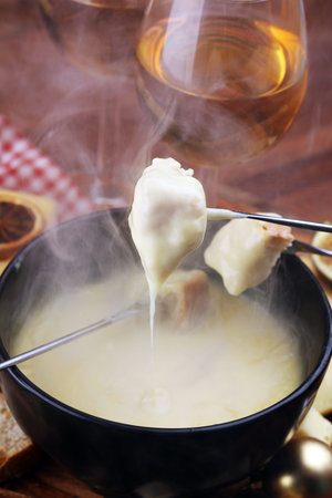 Gourmet Swiss fondue dinner on a winter evening with assorted cheeses on a board alongside a heated pot of cheese fondue with two forks dipping bread and white wine behind in a tavern or restaurantの写真素材