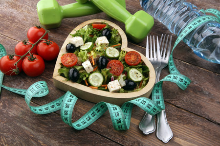 Authentic fresh salad in a wooden heart shaped cup with dumbbells excercise equipment, measuring tape on table. healthy lifestyles, good health Concept with fresh ingredientsの写真素材