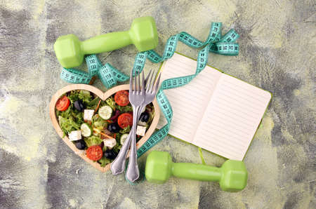Authentic fresh salad in a wooden heart shaped cup with dumbbells excercise equipment, measuring tape on table. healthy lifestyles, good health Concept with fresh ingredientsの写真素材