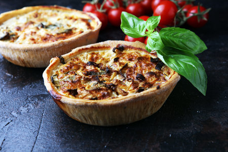 Savory mini quiches or tarts on a rustic board. Flaky dough pies. Fresh basil and tomatoes on...