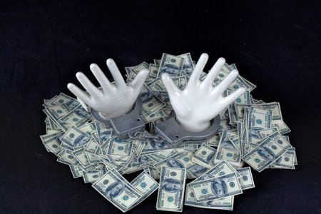 two white ceramic  hands with handcuffs on pile of 100 dollar notes on black background