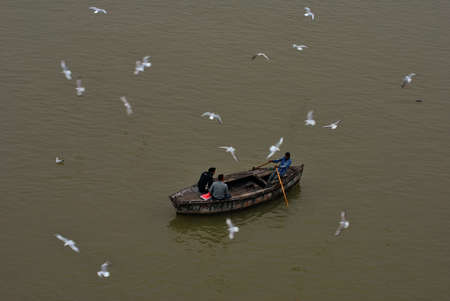 A couple of foreigner tourists enjoying morning boat ride on river Ganges at Varanasi, India. A number of migratory birds are floating and flying on river. Tourist used to feed them. A unique shop variety of items on boat runs by local people is also apprのeditorial素材