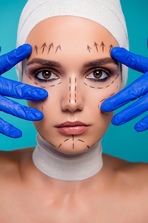 Close up photo of doctor hands in gloves touch girl face with correction marks isolated over cyan backgroundの写真素材