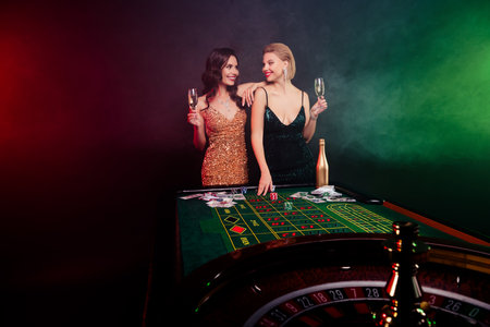 Photo of two stunning partners girls looking each other drinking champagne during blackjack casino over neon backgroundの写真素材