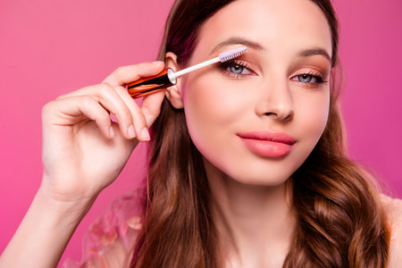 Close up photo of lovely stunning woman applying mascara for extra long lashes isolated on pink color backgroundの写真素材