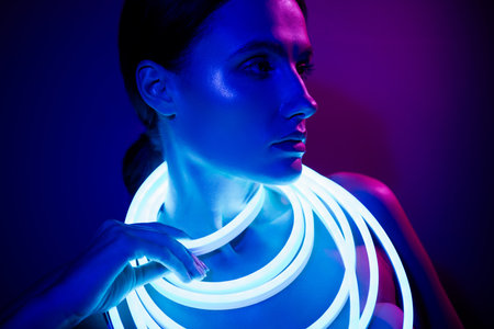 Elegant young woman posing with glowing neon strips against dark blue backgroundの写真素材