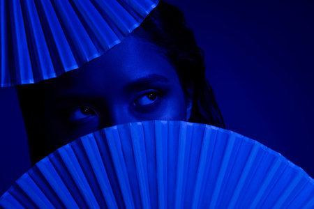 Photo of woman with paper fans close to face looking at empty space with dark blue color background