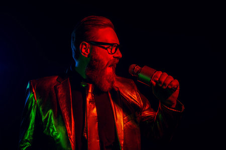 Charismatic man in glossy suit singing passionately against a dark background with neon red and...