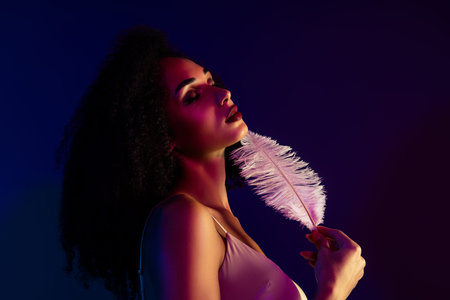 Young woman with curly hair holding pink feather against colorful neon background, promoting style...