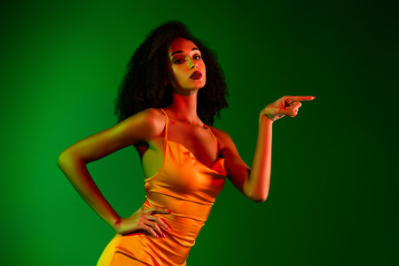 Elegant young woman in a golden cocktail dress posing confidently against a vibrant green gradient backgroundの写真素材