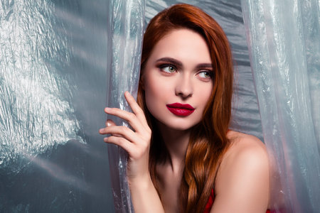 Portrait of a graceful woman with red hair and vibrant makeup posing with a plastic background in an artistic studioの写真素材