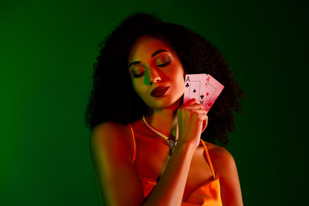 Elegant young woman holding playing cards with confident expression, styled in chic attire against a green backgroundの写真素材
