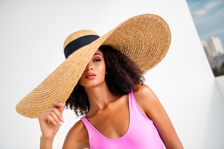 Glamorous young model in oversized straw hat posing confidently in vibrant pink swimwear under daylightの写真素材