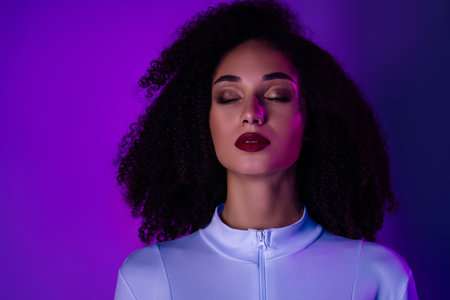 Stylish young woman in fashionable outfit posing with closed eyes under vibrant purple neon lightingの写真素材