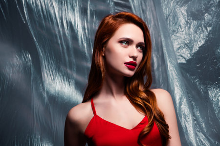 Portrait of a gorgeous woman with red hair in a stylish red dress posing against a plastic backgroundの写真素材