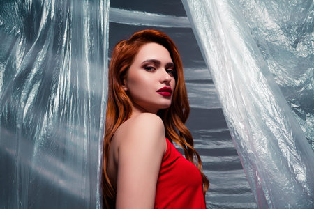Stylish woman with striking red hair in a vibrant red dress, posing gracefully against a dramatic cellophane backdropの写真素材