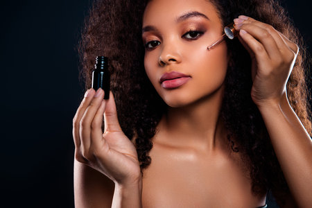 Beautiful woman with curly hair applying serum for skincare and beauty against dark background in black outfitの写真素材