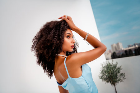 Young woman in a chic blue satin dress posing outdoors with vibrant cityscape in the backgroundの写真素材
