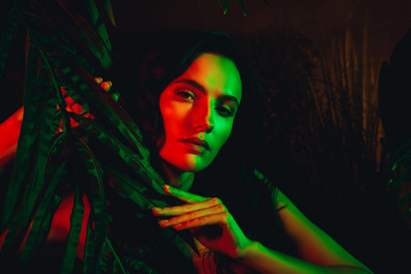 Portrait of a Young Woman Illuminated by Artistic Neon Lighting While Touching Green Tropical Leavesの写真素材