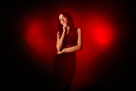 Elegant young woman posing against a vibrant red illuminated smoky background in a studio photoshootの写真素材