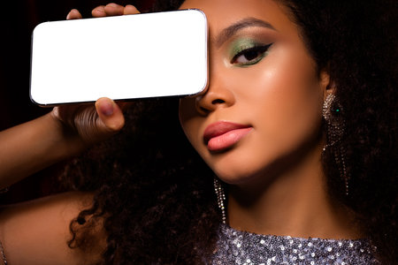 Stylish woman posing holding smartphone with a neutral screen beside her face, showcasing makeup and luxurious stylingの写真素材