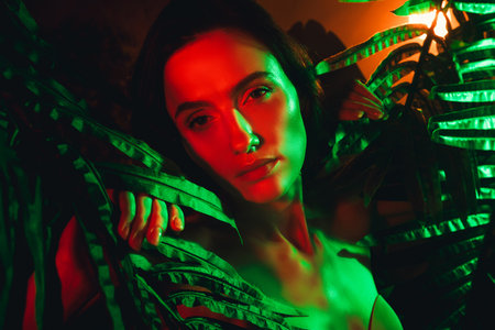 Beautiful young woman surrounded by tropical leaves under dramatic red and green neon lighting, evoking exotic vibes.の写真素材