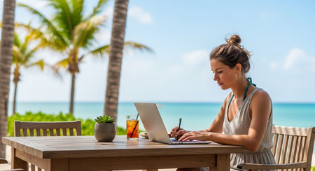 woman works on her laptop at a table outside with a beautiful ocean view and palm trees on a sunny dayの素材