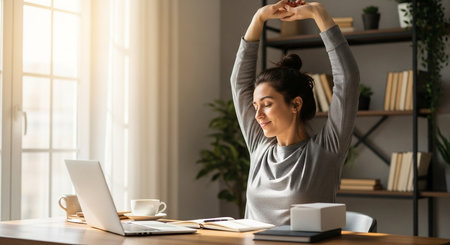 a woman is stretching at her desk with a laptop, books, and coffee in a bright and airy home office spaceの素材