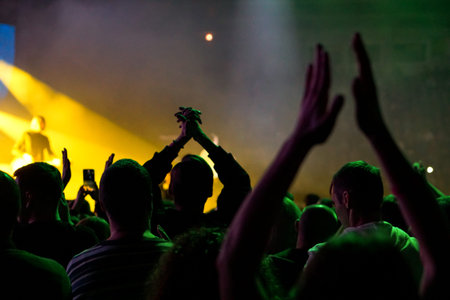 Audience with hands raised at a music festival and lights streaming down from above the stage. Soft...