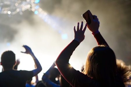 Audience with hands raised at a music festival and lights streaming down from above the stage. Soft...