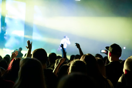 Audience with hands raised at a music festival and lights streaming down from above the stage. Soft...
