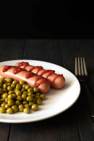 Sausages with ketchup, a side dish of peas on a dark background. Front view. White plate and stainless steel fork.の写真素材
