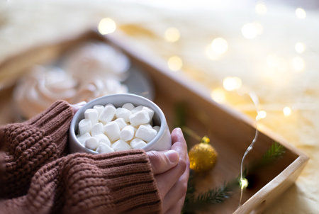 A large mug with cocoa in the hands of a girl in a sweater, in the background of a meringue and...