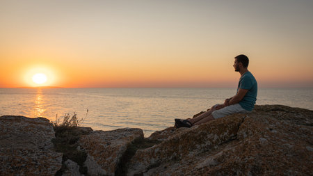 A man enjoys the view of the sunset on the sea, sitting on a rock. Side view, silhouette.