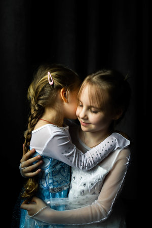 Two younger sisters hug, photos in costumes on a dark background. Front view.