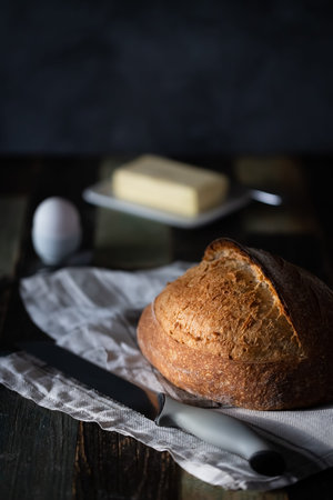 Homemade sourdough wheat bread on a dark background with an egg in a ceramic stand and butter, next to a bread knife. Dark background, a wooden table.の写真素材